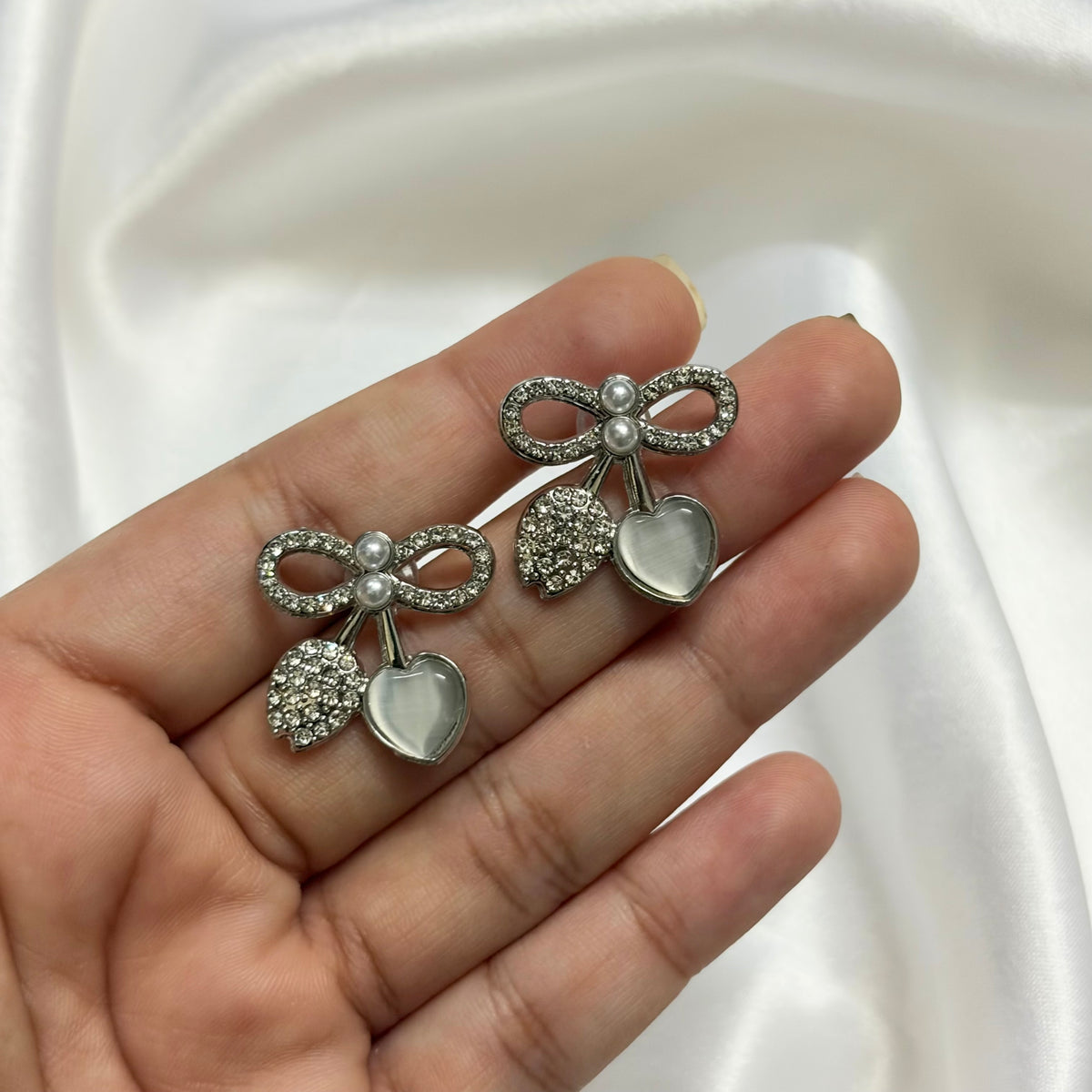 A pair of silver-toned bow and heart stud earrings with clear rhinestones and milky white heart charms, held on a person's fingers against a draped white satin background.