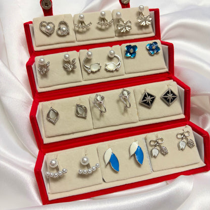 A red jewelry display box filled with multiple pairs of earrings and rings, arranged on cream velvet compartments against a white satin fabric background.