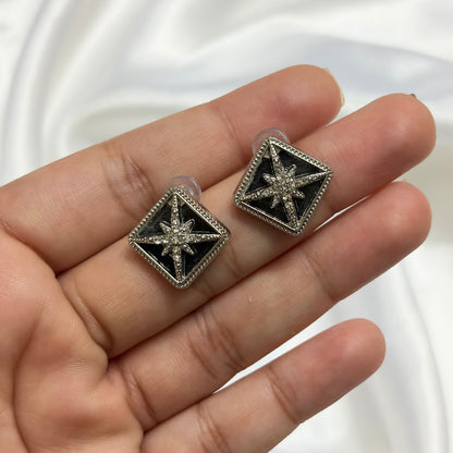 A pair of silver-toned square stud earrings with black enamel centers and starburst details, shown resting on a person's hand against a white satin background.