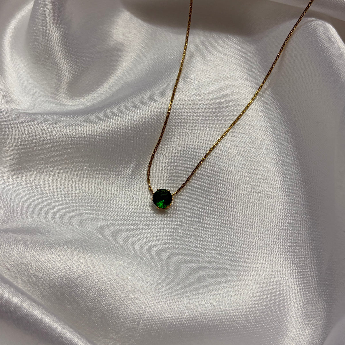 A delicate gold chain necklace with a small round black pendant featuring a vibrant green center, displayed on draped white satin fabric.