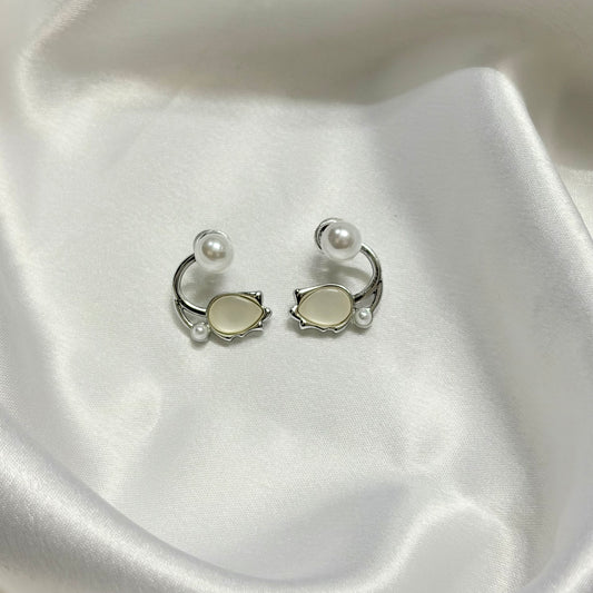 A pair of silver-toned C-shaped hoop earrings with oval cream-colored stones and small pearl-like beads, displayed on draped white satin fabric.