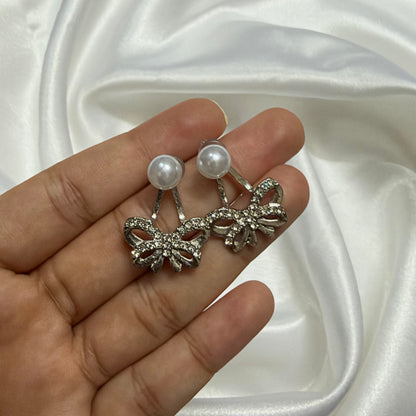 A pair of silver-toned bow earrings with white pearl studs, held on a person's fingers against a draped white satin fabric background.
