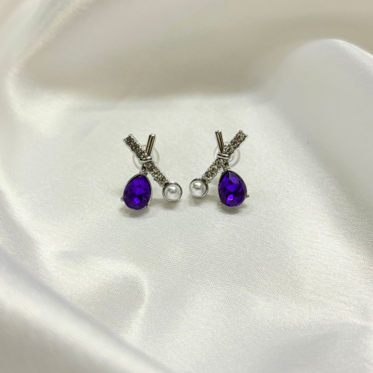A pair of silver stud earrings with teardrop-shaped purple gemstones, a small silver bead, and a clear round pearl, displayed on a white satin fabric background.