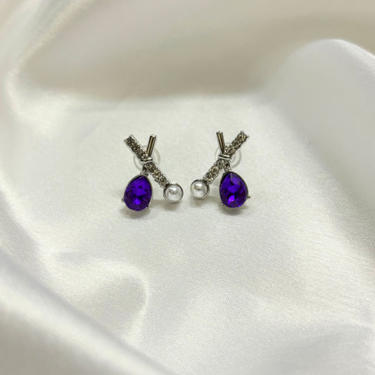 A pair of silver stud earrings with teardrop-shaped purple gemstones, a small silver bead, and a clear round pearl, displayed on a white satin fabric background.
