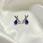 A pair of silver stud earrings with teardrop-shaped purple gemstones, a small silver bead, and a clear round pearl, displayed on a white satin fabric background.