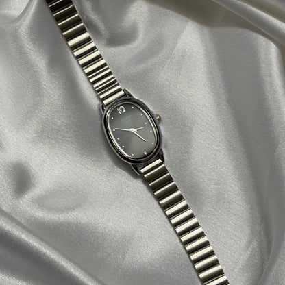 A silver-toned oval watch with a matching metal link bracelet, displayed diagonally on draped satin fabric.