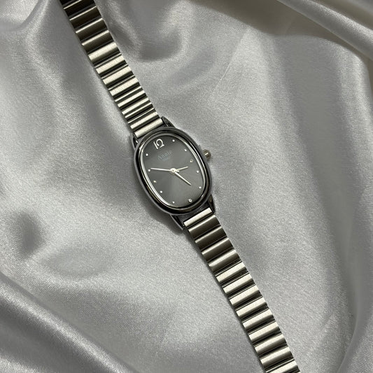 A silver-toned oval watch with a matching metal link bracelet, displayed diagonally on draped satin fabric.