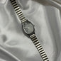 A silver-toned oval watch with a matching metal link bracelet, displayed diagonally on draped satin fabric.