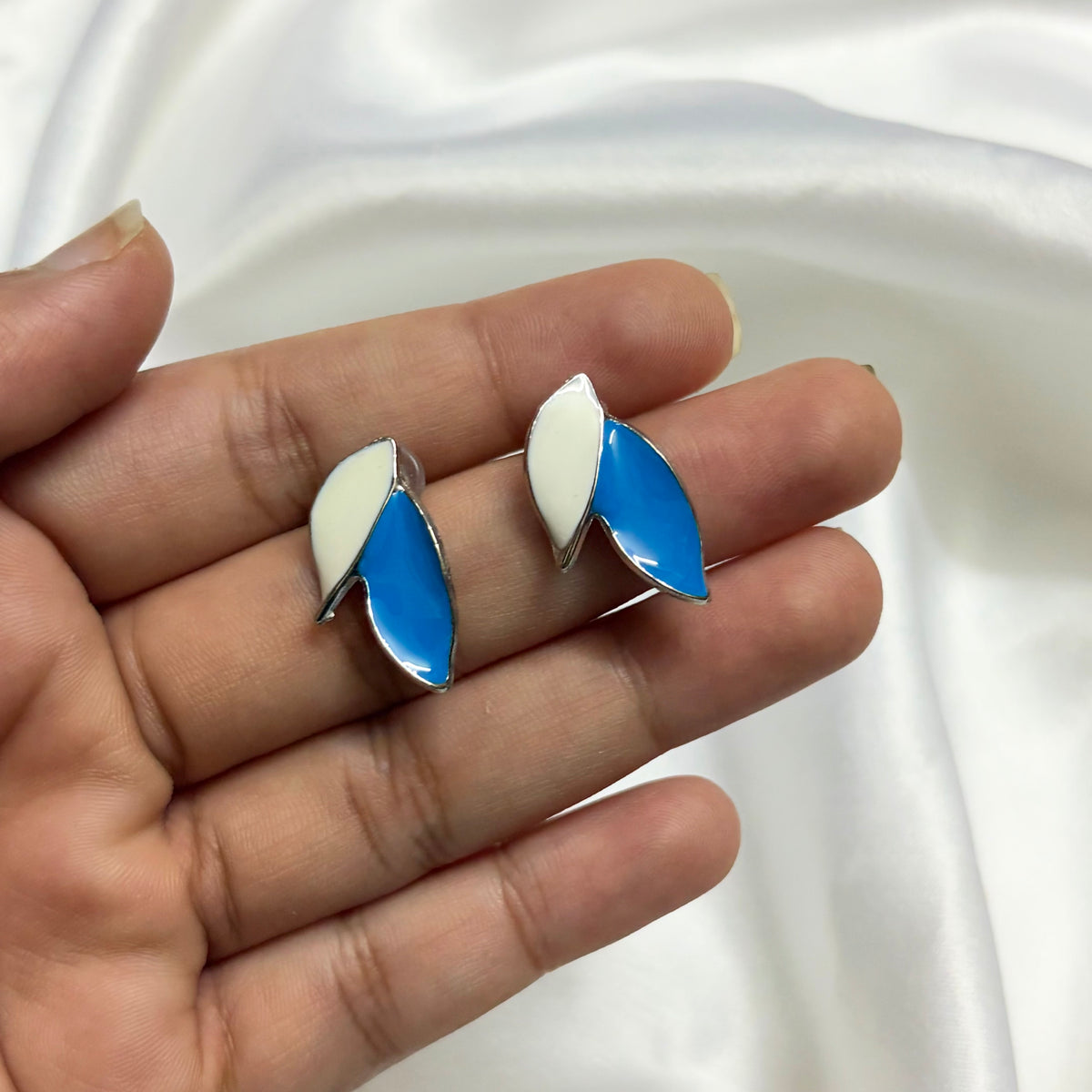 A pair of silver-tone stud earrings with white and blue enamel leaf-like shapes, held in a person's hand against a white satin fabric background.