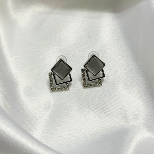 A pair of geometric stud earrings with layered square and rectangular silver-tone frames, one featuring a smooth dark gray inset and the other accented with small clear stones, displayed on a white satin fabric background.