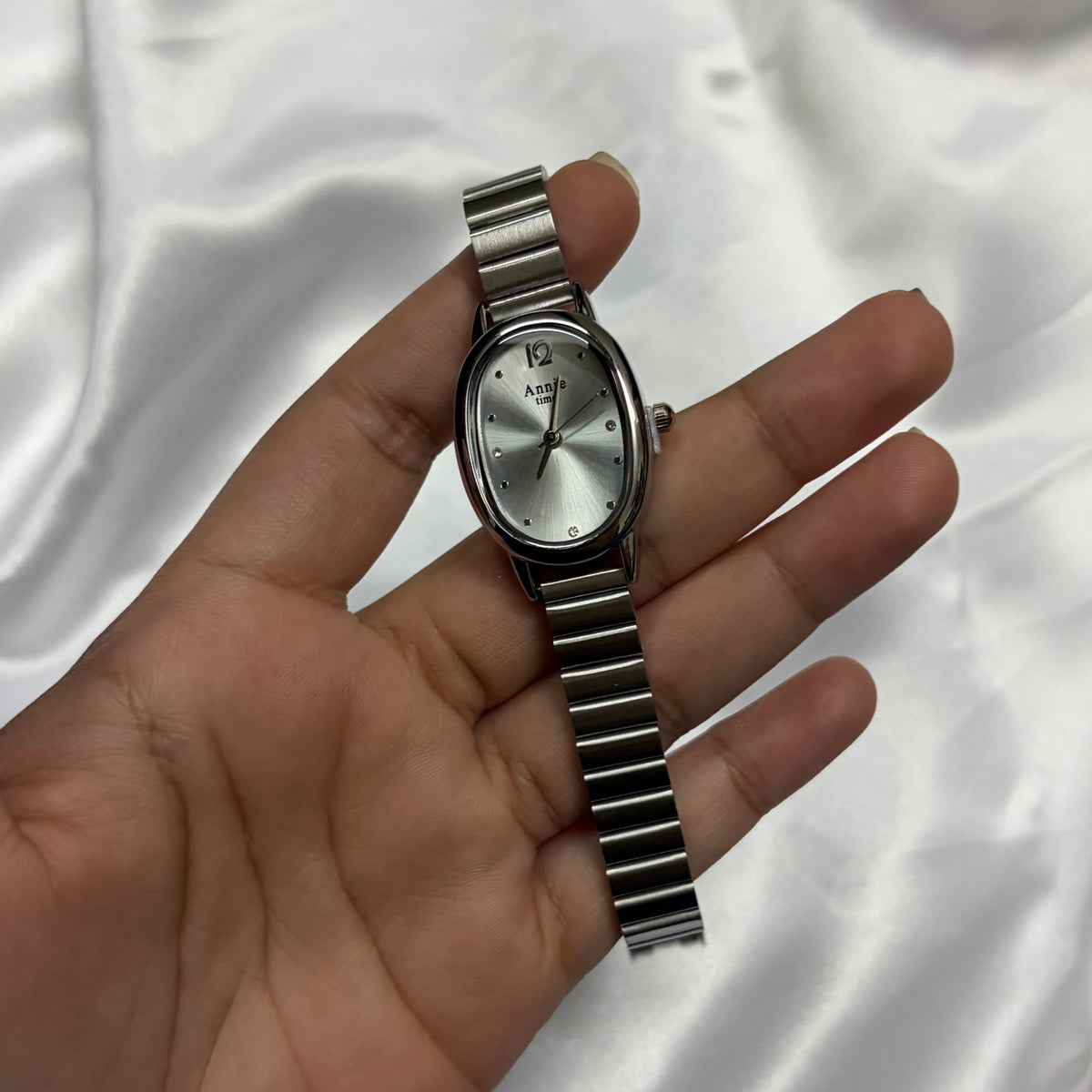 A silver-tone women's wristwatch with an oval silver dial and matching metal link bracelet, held in a person's hand against a white satin fabric background.