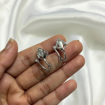 A pair of silver-toned floral hoop earrings with clear marquise-cut stones and small accent stones, shown resting on a person's fingers against a white satin fabric background.