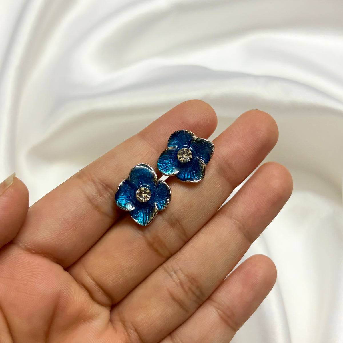 A pair of blue flower-shaped stud earrings with silver-toned metal edges and clear rhinestone centers, held on a person's hand against a white satin fabric background.