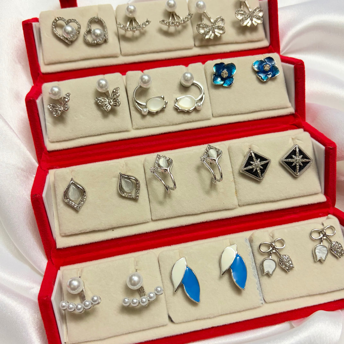 A red jewelry display box filled with multiple pairs of earrings, including silver-tone designs with pearls, flowers, leaves, and geometric motifs, set against a white satin background.