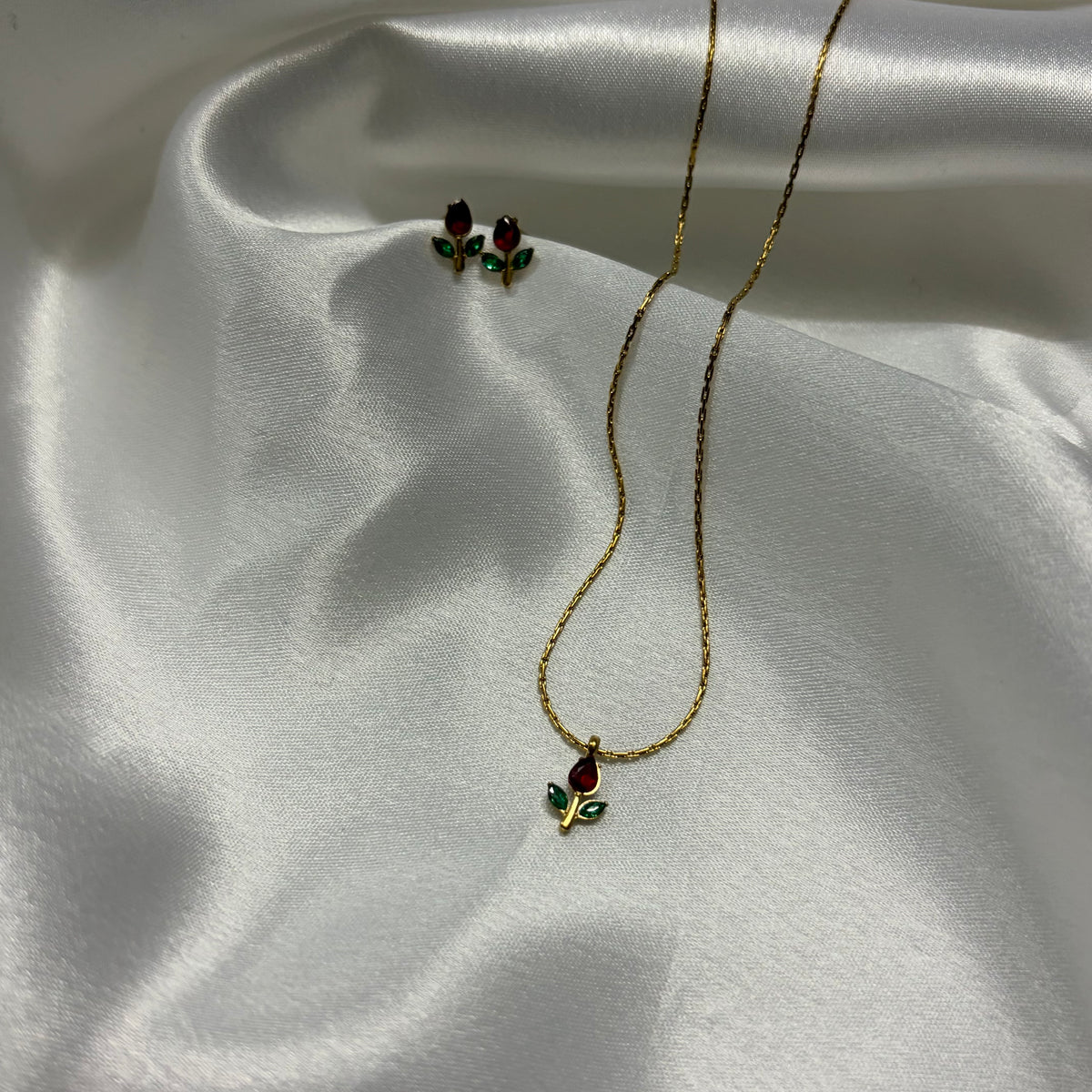 A gold necklace and matching stud earrings with small red rose-shaped stones and green leaf accents, displayed on draped white satin fabric.