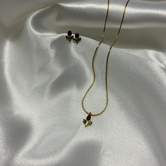 A gold necklace and matching stud earrings with small red rose-shaped stones and green leaf accents, displayed on draped white satin fabric.